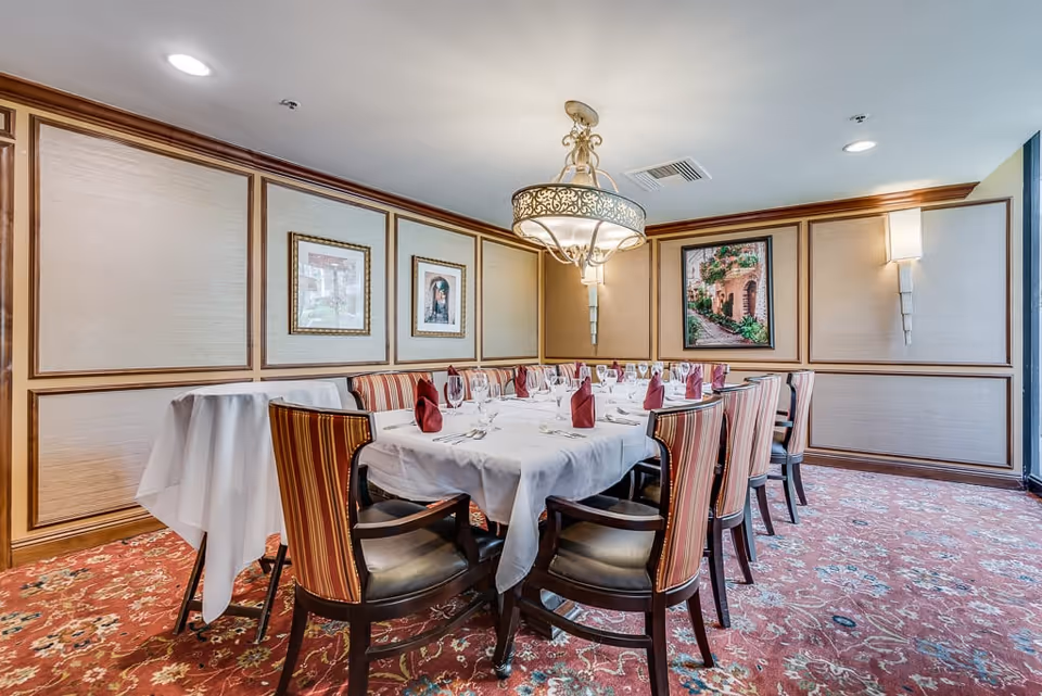A formal dining room with a long rectangular table covered with a white tablecloth, set with wine glasses, napkins folded in a standing style, and silverware. The room features striped upholstered chairs, framed artwork on the walls, a decorative chandelier hanging from the ceiling, and patterned carpet flooring.
