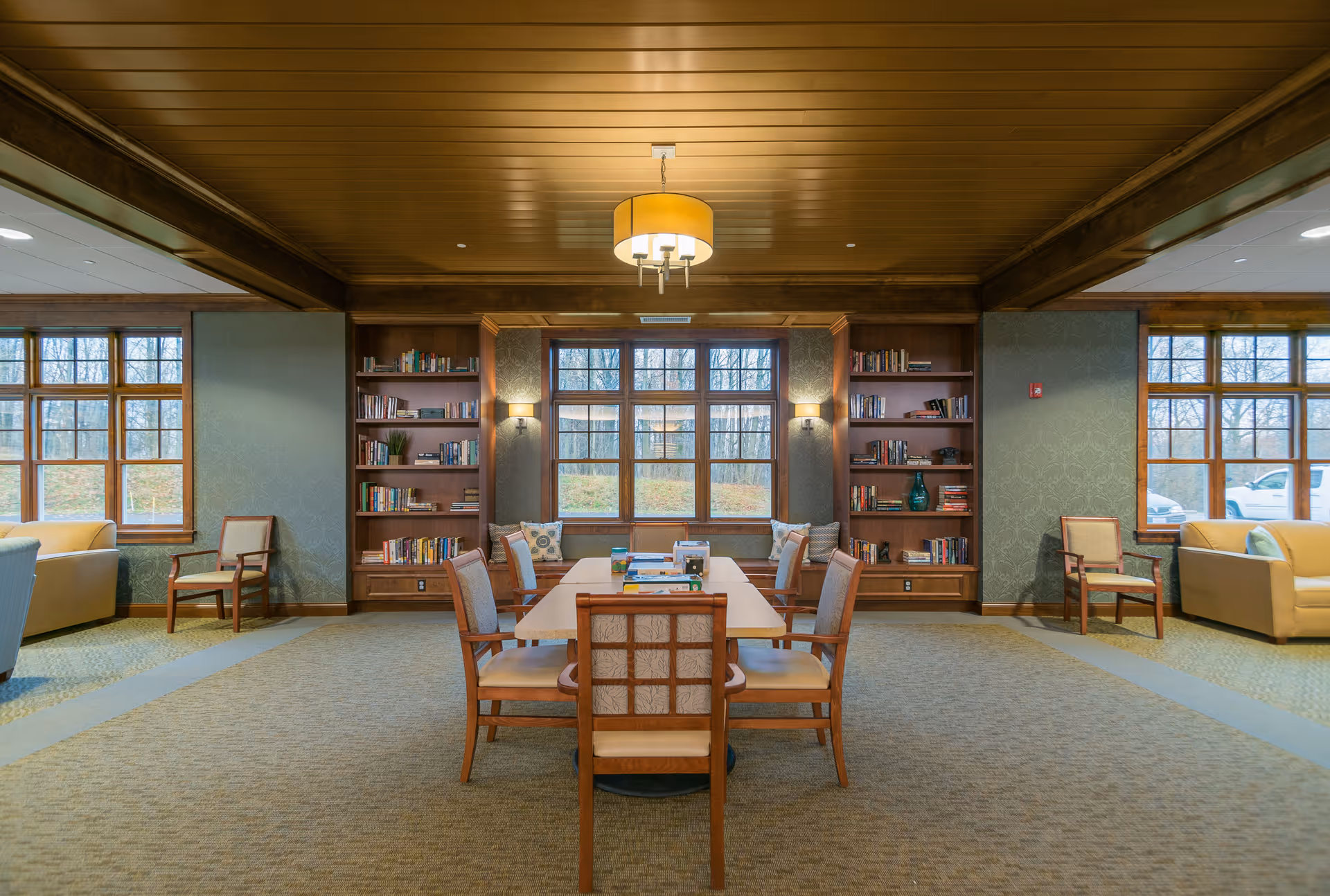 A cozy and well-lit common area in Armstrong Memory Care Assisted Living featuring a central wooden table surrounded by six cushioned chairs. The room has large windows with views of trees outside, built-in wooden bookshelves filled with books and decorative items, two armchairs near the windows, and a warm wooden ceiling with a modern hanging light fixture.