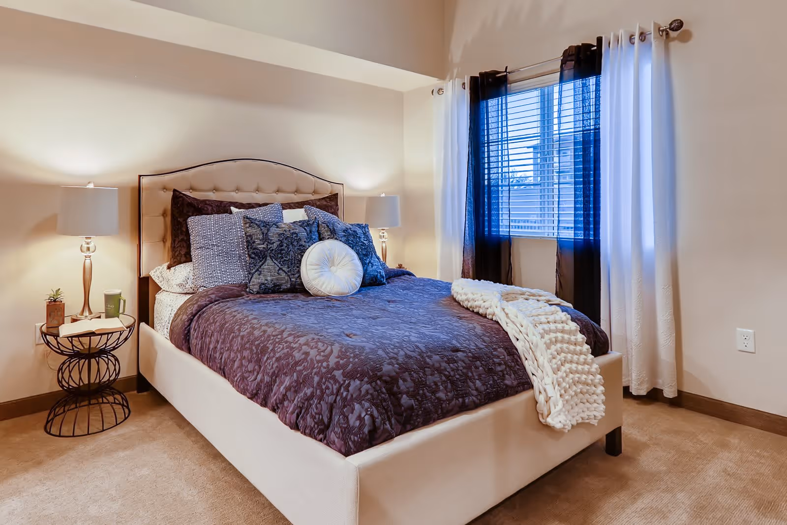 A cozy bedroom with a beige upholstered bed frame and headboard, dressed in dark purple bedding with multiple decorative pillows and a white knitted throw blanket. There are two bedside tables with matching lamps on either side of the bed. A window with white and dark curtains is on the right wall, allowing natural light into the room.