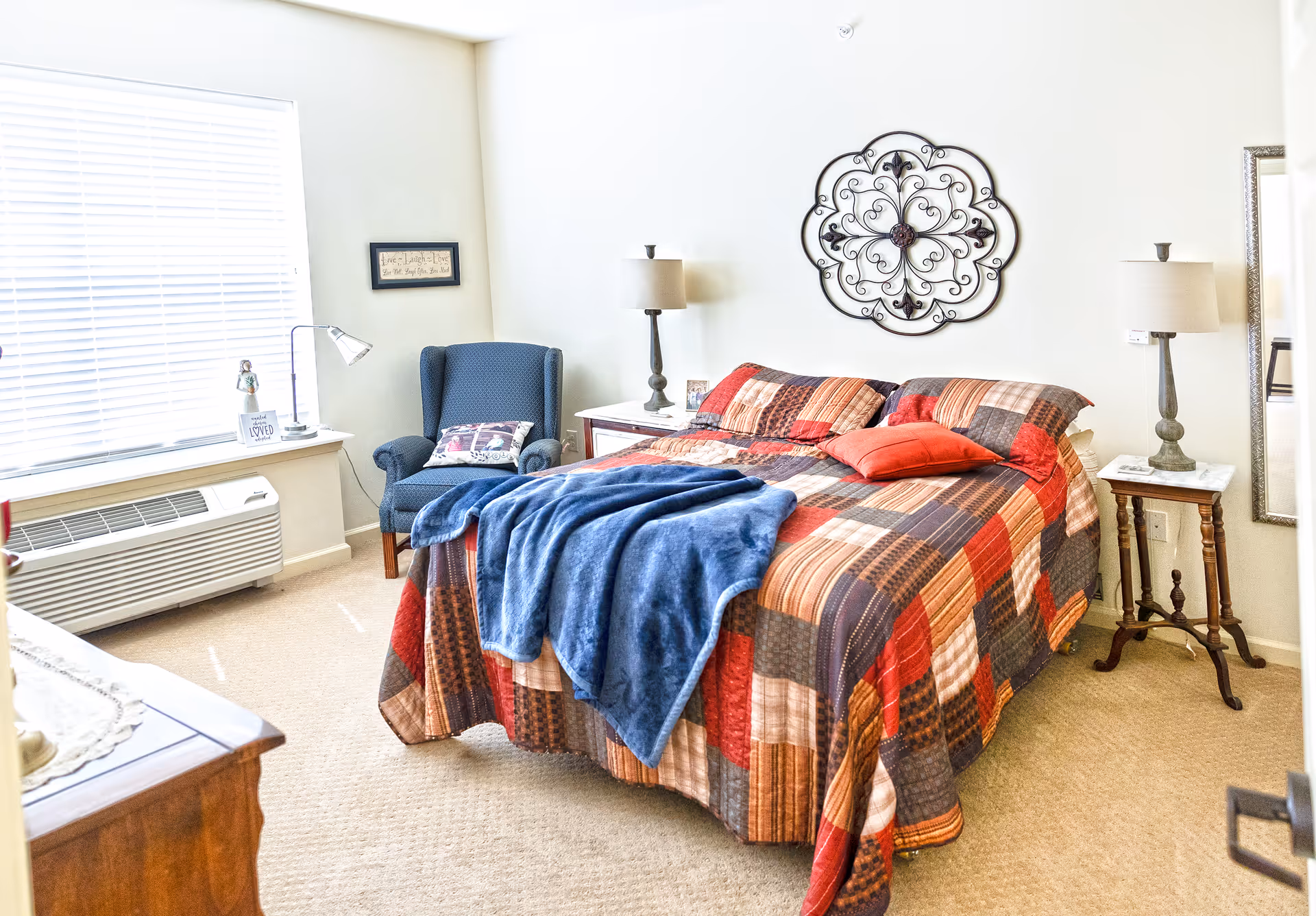 A cozy bedroom with a large bed covered in a patchwork quilt with red, brown, and beige tones, and a blue blanket draped over it. There are two bedside tables with matching lamps, a blue upholstered armchair with a decorative pillow, a window with blinds, and a decorative metal wall art above the bed.