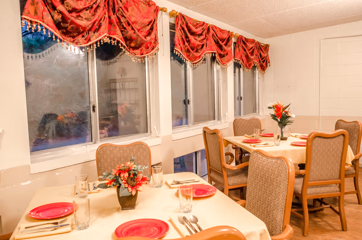 Dining room with tables covered in beige tablecloths, each set with red plates, glasses, and silverware. The room has upholstered chairs with wooden frames and windows adorned with red valance curtains featuring gold trim and tassels. Floral centerpieces decorate the tables.