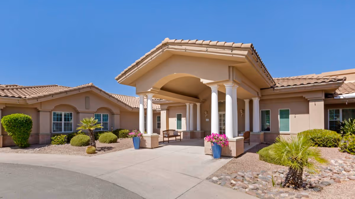 Exterior view of Brookdale Oro Valley senior living facility entrance with a covered porch supported by white columns, benches, potted flowers, and desert landscaping including small palm trees and bushes under a clear blue sky.