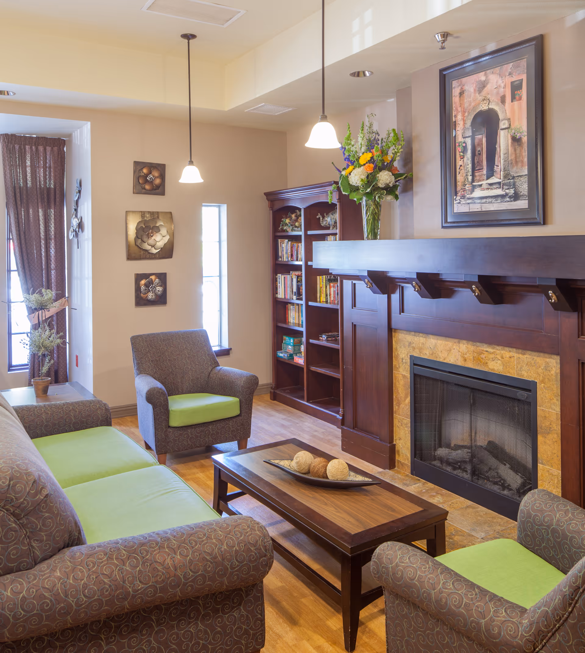 A cozy living room with a patterned sofa and two armchairs with green cushions, a wooden coffee table with decorative balls, a fireplace with a wooden mantle, a bookshelf filled with books, a vase with colorful flowers on the mantle, and wall art above the fireplace and on the adjacent wall. The room has wooden flooring and pendant lights hanging from the ceiling.
