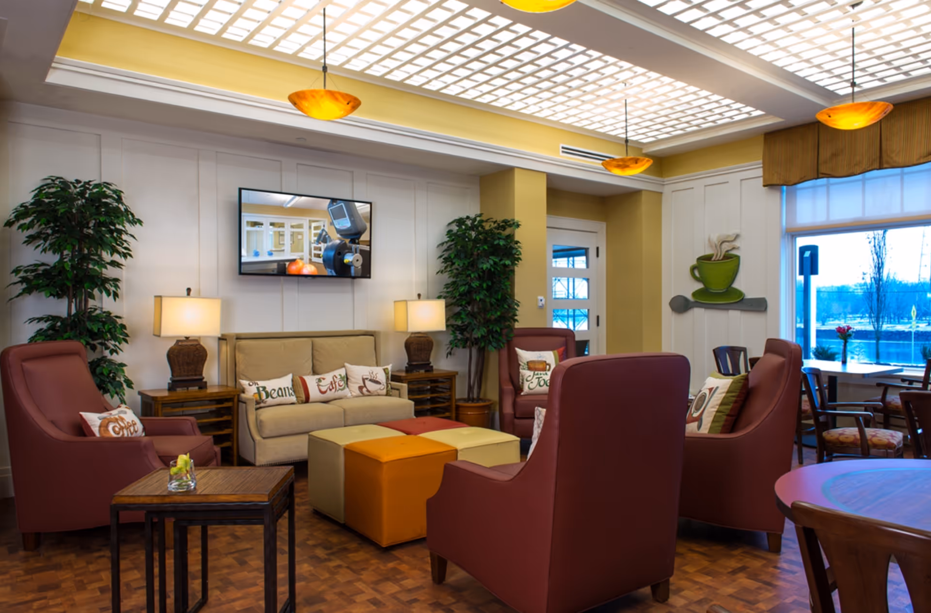 A cozy and well-lit living room area with a beige sofa adorned with decorative pillows, four maroon armchairs, two wooden side tables with lamps, and two potted plants. The ceiling features a grid pattern with recessed lighting and three hanging amber pendant lights. A flat-screen TV is mounted on the wall above the sofa. There is a large window with a view of the outdoors and a wall decoration of a green coffee cup with steam and a spoon.
