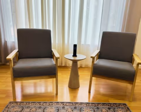 Two gray cushioned armchairs with wooden frames positioned on either side of a small round wooden side table holding a remote control, set in front of sheer white curtains with a wooden floor and a patterned rug partially visible at the bottom.