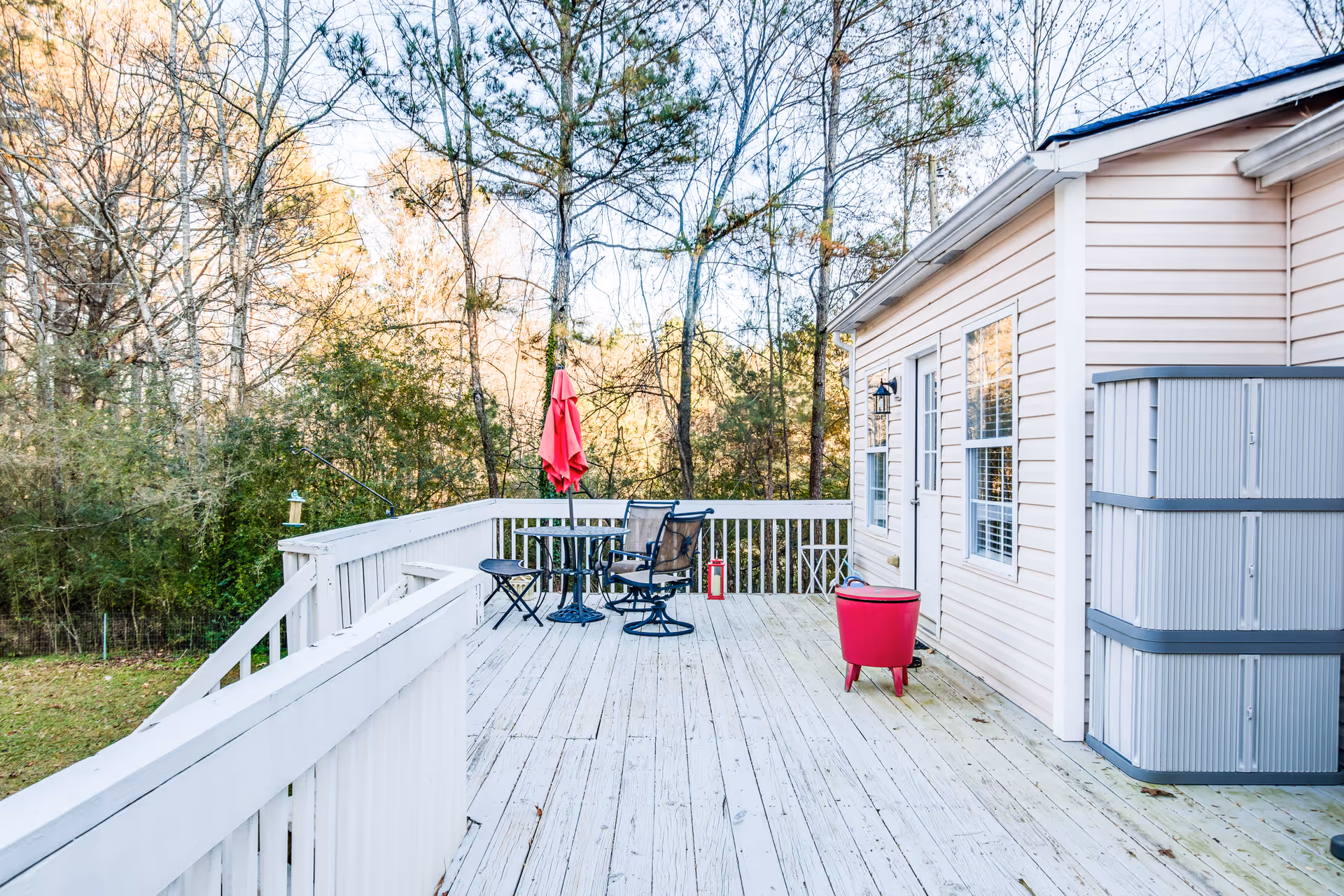 A spacious wooden deck attached to a light-colored residential building, featuring a small round table with two chairs and a closed red umbrella. There is a red cooler and a large gray storage container on the deck. Trees and greenery surround the area in the background.