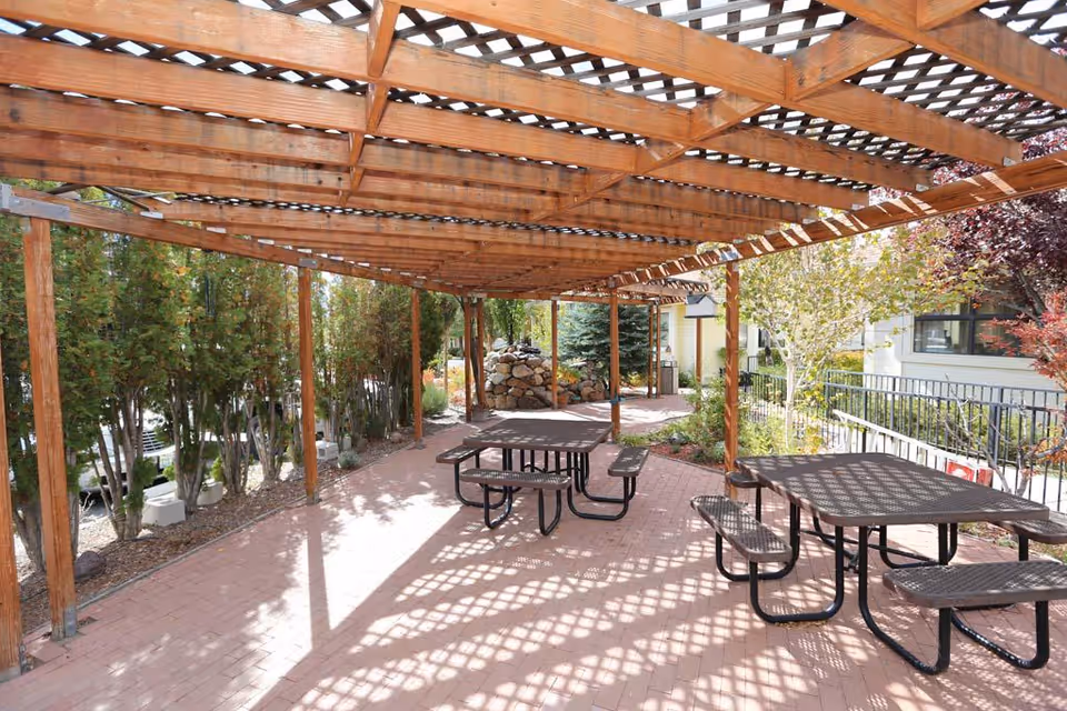Wooden pergola-covered outdoor seating area with picnic tables on brick pavers surrounded by trees and landscaping.