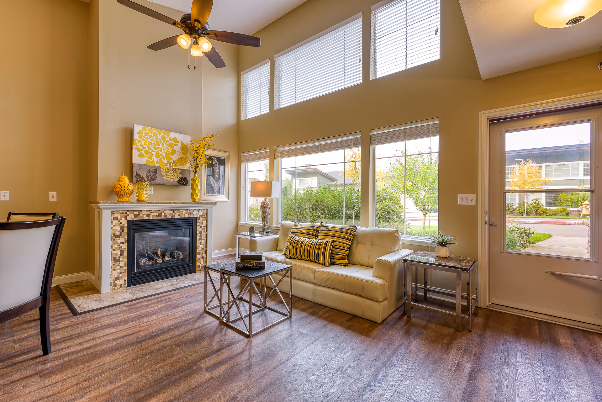 A bright living room with a white leather sofa adorned with yellow and striped pillows, a modern glass coffee table, and a fireplace with a mosaic tile surround. Large windows let in natural light and offer a view of greenery outside. The room features wood flooring, a ceiling fan with lights, and decorative yellow vases and artwork above the fireplace.