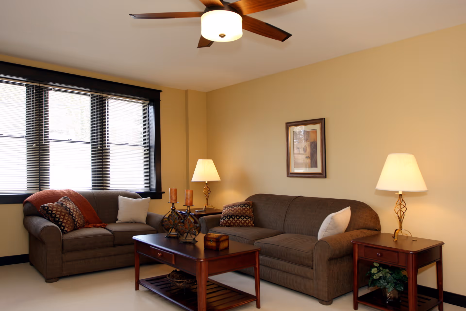 A cozy living room with two brown sofas adorned with decorative pillows and a throw blanket. A wooden coffee table with candles and a small box sits between the sofas. Two side tables with lamps flank the sofas. A ceiling fan with a light fixture is mounted on the ceiling, and a framed picture hangs on the beige wall. A large window with blinds lets in natural light.