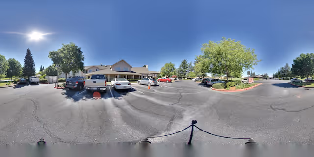 Panoramic view of a senior living facility parking lot and building entrance with parked cars, trees, and a clear blue sky.