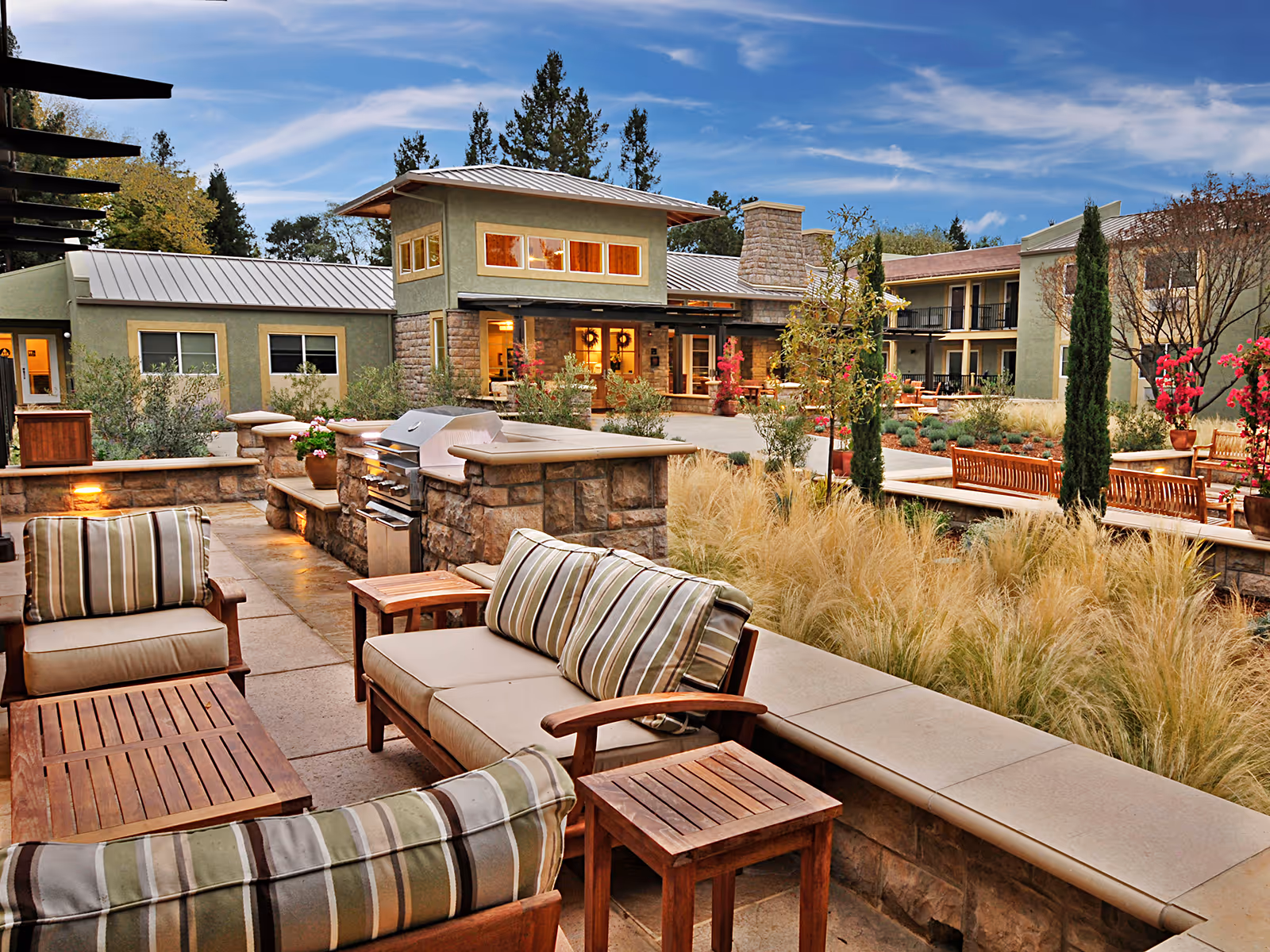 Outdoor patio area at Atria Tamalpais Creek featuring cushioned wooden seating with striped pillows, a built-in stone grill, landscaped garden with ornamental grasses and flowering plants, and a multi-story building in the background under a partly cloudy sky.