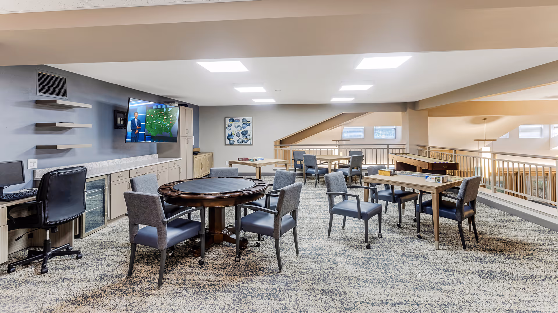 A spacious common area in a senior living facility with multiple tables and chairs arranged for group activities. There is a round table with six chairs in the foreground, a rectangular table with four chairs to the right, and another table with chairs in the background near a railing overlooking a lower level. A flat-screen TV mounted on the wall displays a weather forecast. The room has bright overhead lighting, carpeted floors, and neutral-colored walls with some decorative artwork.