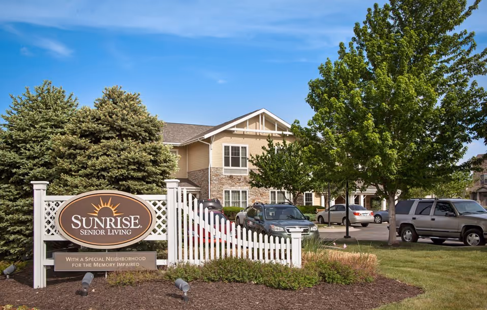 Exterior view of Sunrise Senior Living facility with a white picket fence and a sign in front. The building is two stories with beige siding and stone accents. Several cars are parked in the parking lot, and there are green trees and shrubs around the area under a clear blue sky.