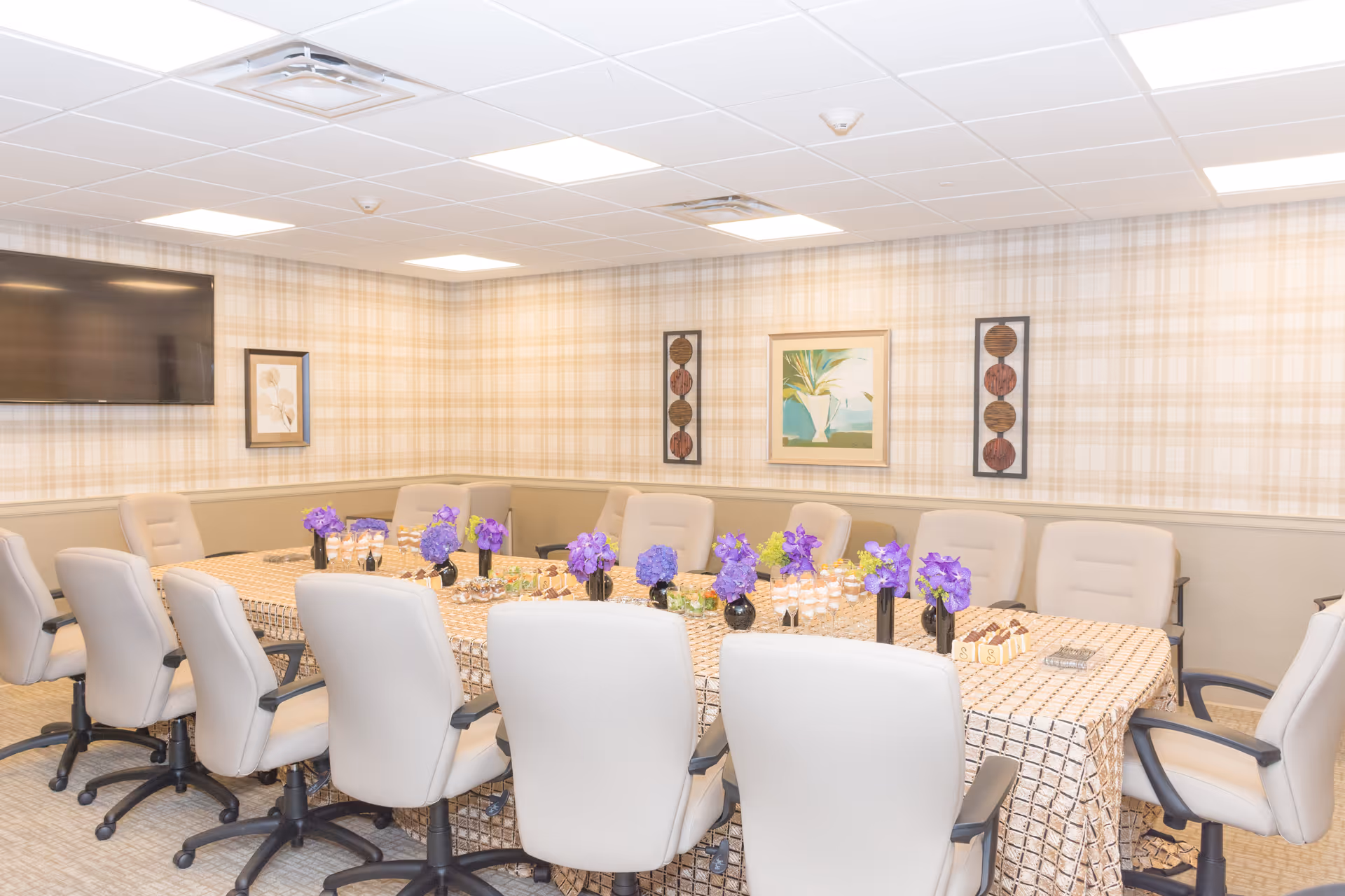 A conference or meeting room with a long rectangular table covered with a patterned tablecloth. The table is surrounded by beige office chairs on wheels. The table is decorated with small vases of purple flowers and some small desserts or snacks. The walls have a plaid wallpaper with framed artwork, and a large flat-screen TV is mounted on one wall. The ceiling has recessed lighting panels.