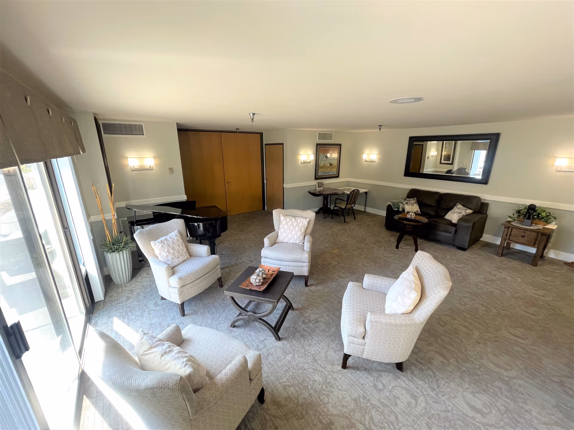 A spacious senior living common area with four upholstered armchairs arranged around a small coffee table. There is a black grand piano near the back wall, a small table with two chairs, a dark brown sofa with decorative pillows, a large mirror on the wall, and a side table with a plant and decorative items. The room has carpeted flooring, light-colored walls, and large windows letting in natural light.