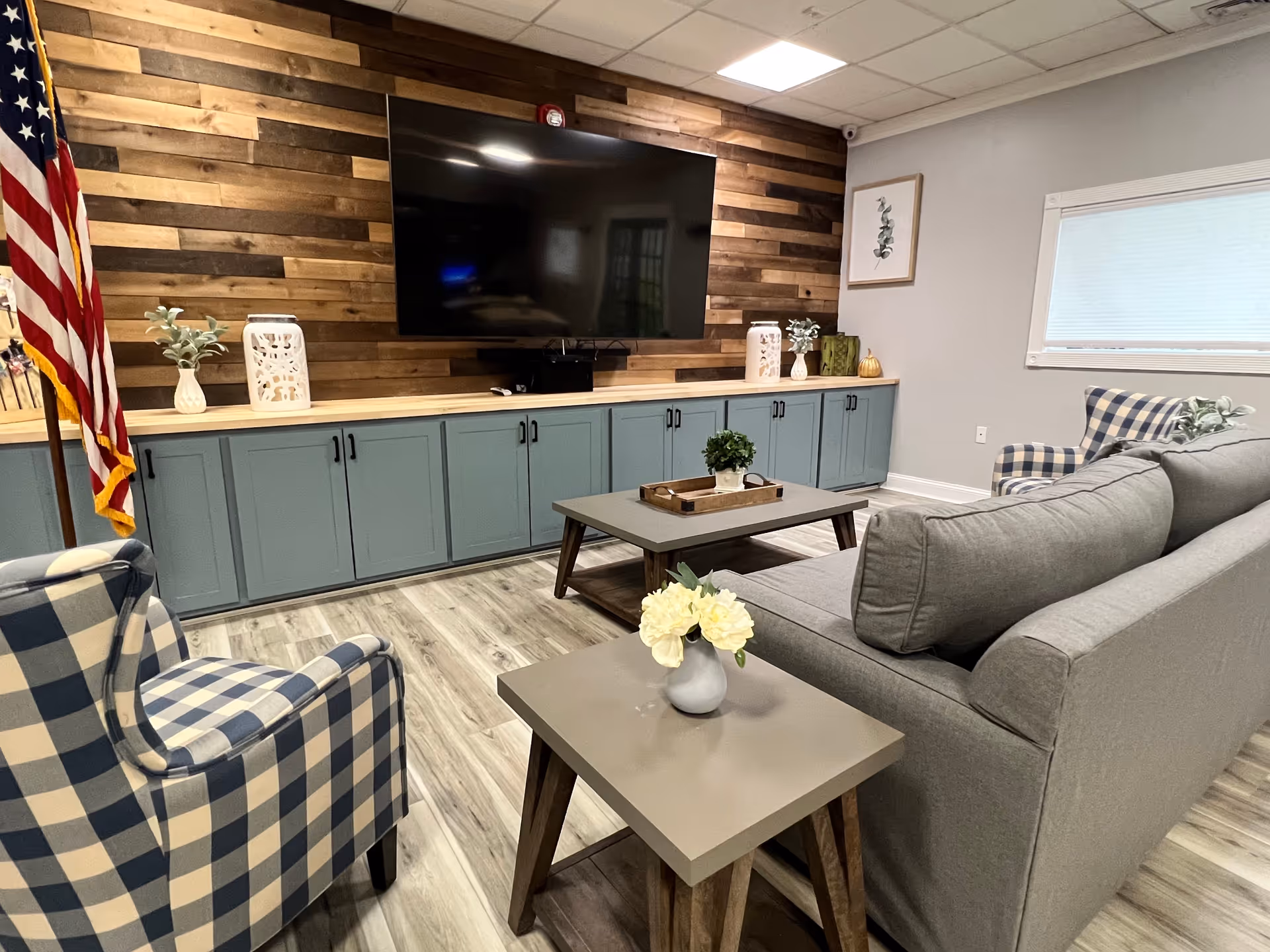 A cozy living room with a large flat-screen TV mounted on a wooden accent wall. Below the TV is a long cabinet with blue doors and black handles. The room features a gray sofa, a blue and white checkered armchair, a coffee table with a small plant, and a side table with a vase of white flowers. An American flag stands in the corner, and a window with a white blind is on the right wall.