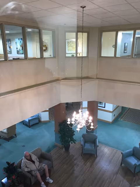 View from an upper floor overlooking a common area in a senior living facility. The area has blue carpet, wooden flooring, several armchairs, a potted plant, and a chandelier hanging from the ceiling. A man is sitting on one of the armchairs. The upper floor has windows looking into rooms or offices.