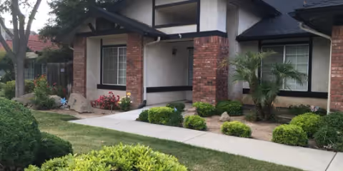 Front exterior view of a single-story residential building with brick and white walls, a covered entrance, and a well-maintained garden with bushes, small trees, and a concrete walkway.