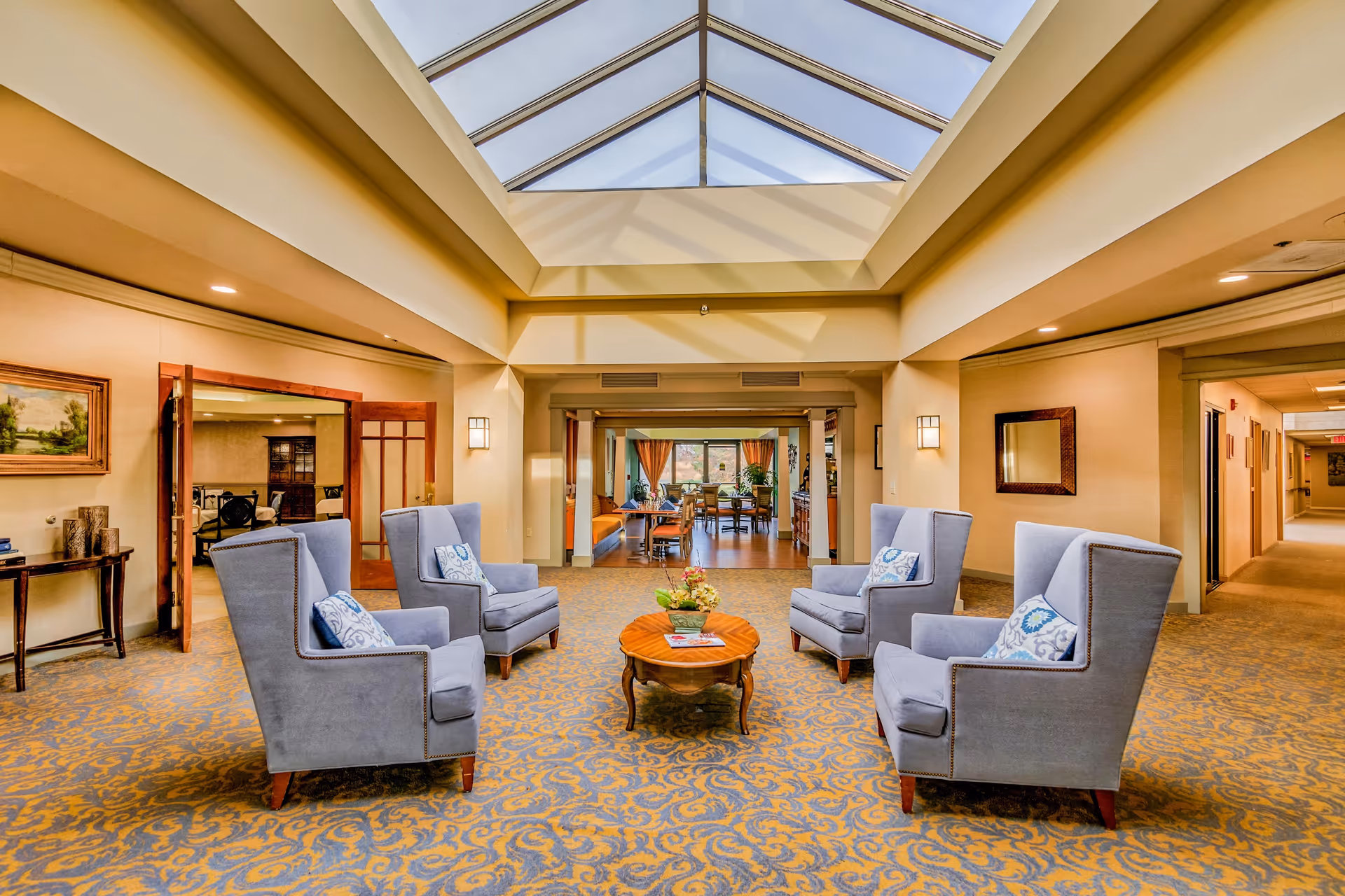 A bright and spacious common area in a senior living facility with four blue upholstered armchairs arranged around a wooden coffee table. The room features a large skylight ceiling allowing natural light to fill the space. There is patterned carpet flooring and warm beige walls with wall sconces. In the background, there is another seating area with orange chairs and large windows with curtains.