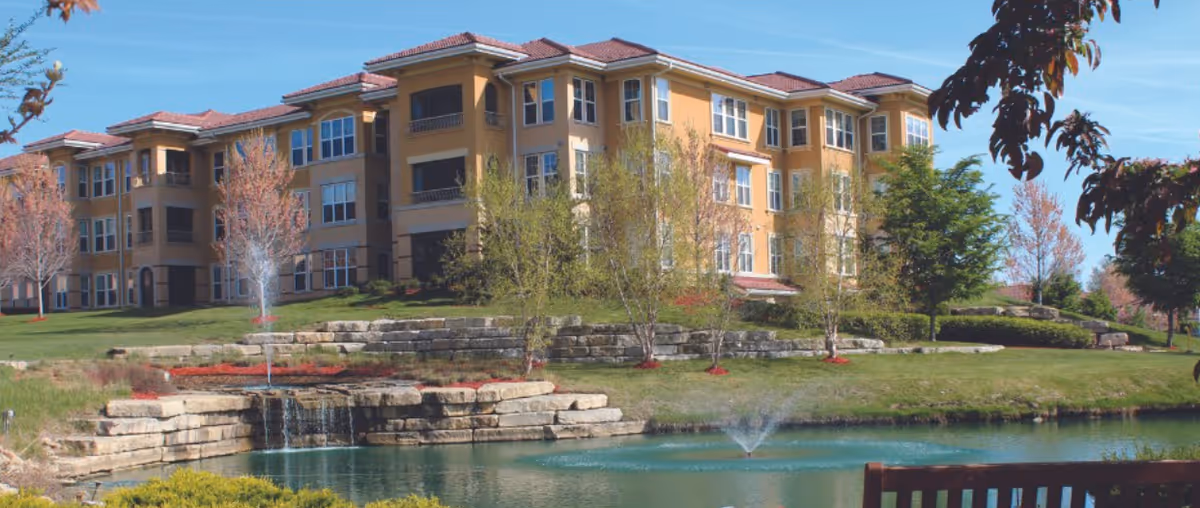 A large multi-story residential building with a yellow exterior and red-tiled roof, surrounded by trees and landscaping. In the foreground, there is a pond with a small waterfall and a fountain, along with a wooden bench partially visible on the right side.