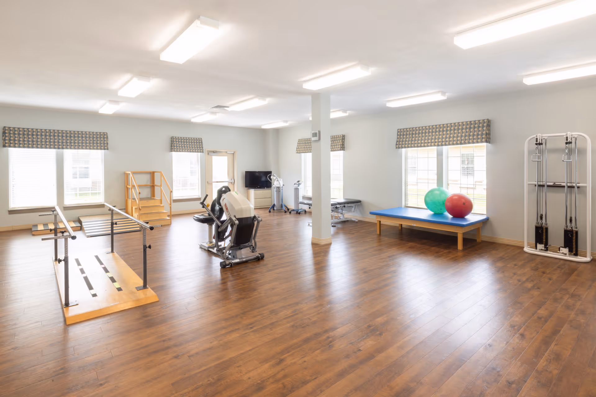 Bright rehabilitation/exercise room with parallel bars, exercise bike, therapy table, and exercise balls