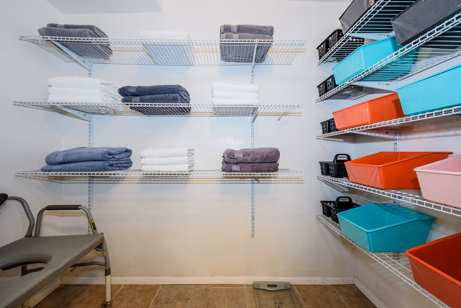 A storage room with white wire shelves holding neatly folded towels in white, gray, and purple colors on the left side, and colorful plastic bins in teal, orange, pink, and black on the right side. A gray shower chair is partially visible on the left, and a digital scale is on the floor.