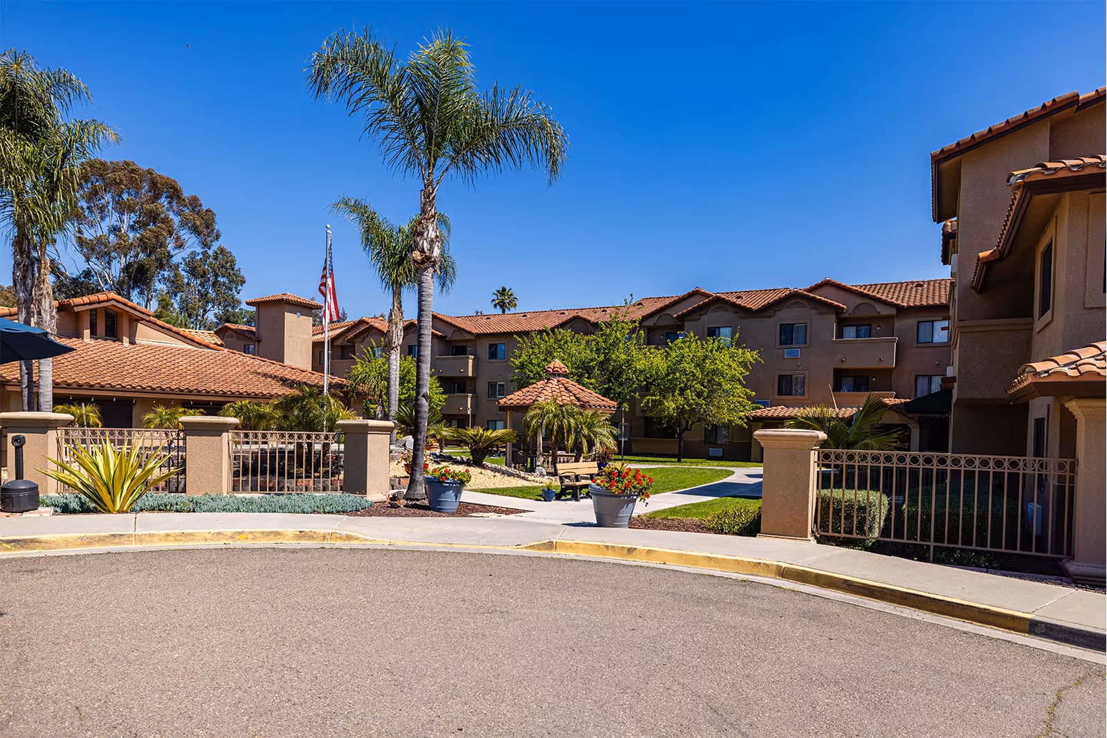 Front courtyard and entrance of a Mediterranean-style retirement community with palm trees, potted plants, and multi-story tan buildings with red tile roofs.