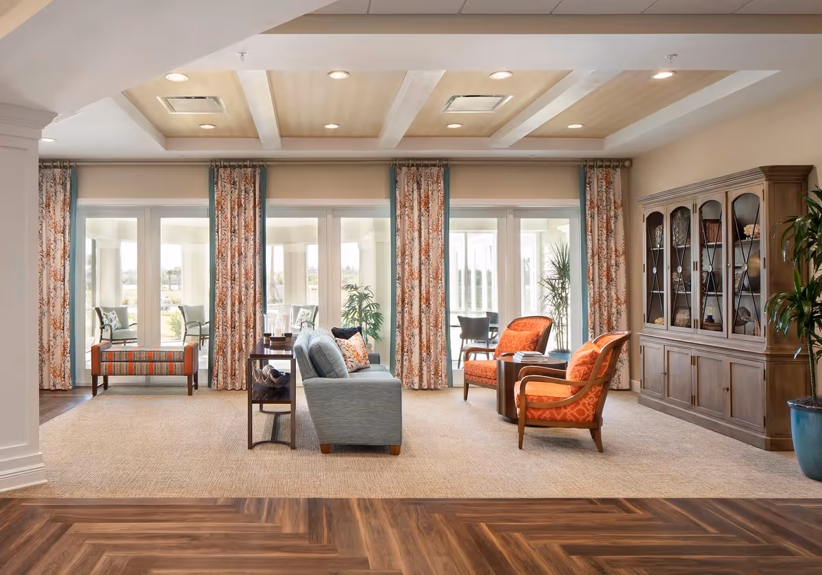 Spacious, well-lit lounge with a sofa, two orange armchairs, patterned curtains and a wooden display cabinet.