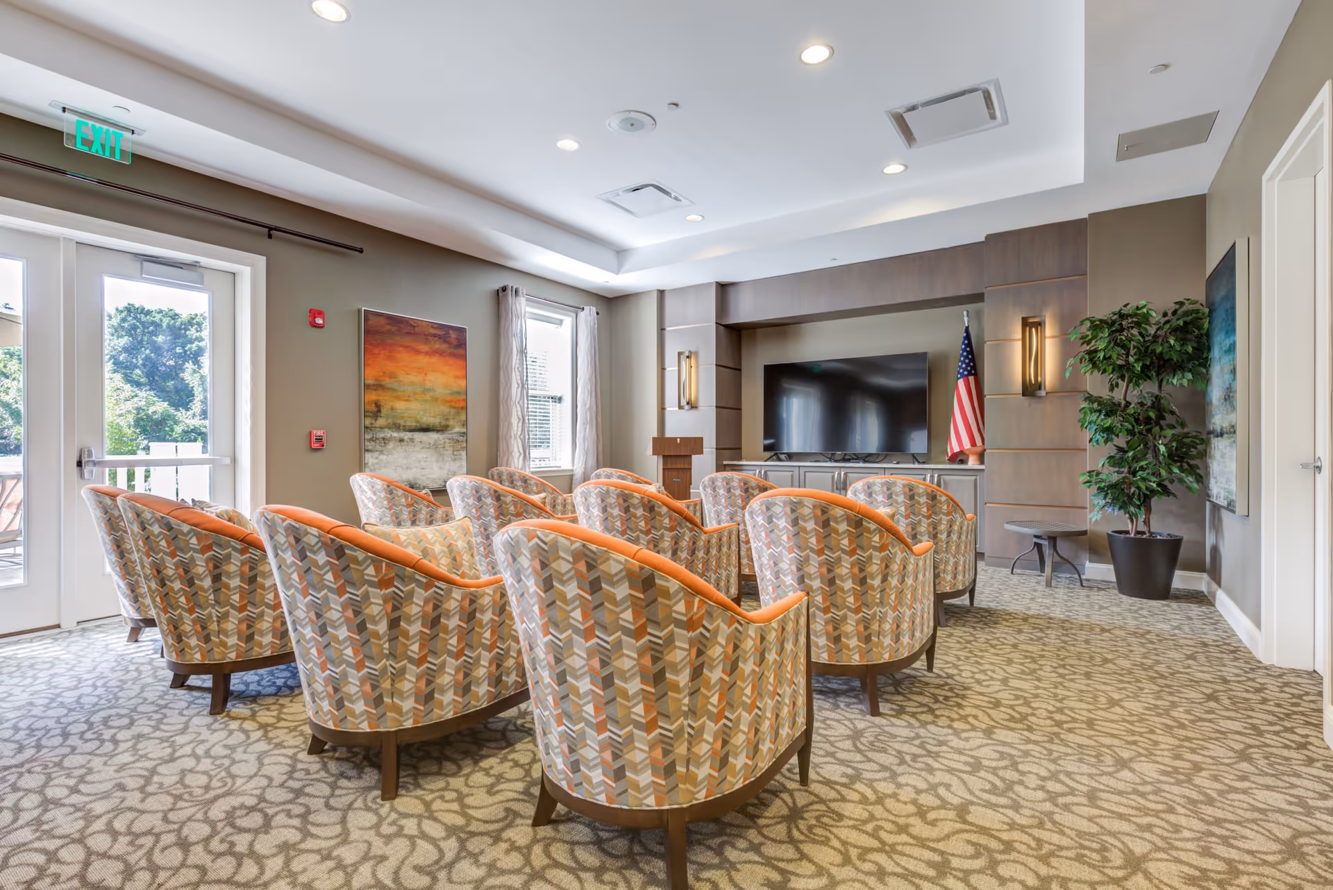 Community media room with rows of patterned armchairs facing a large flat-screen TV and podium, with windows and an American flag.