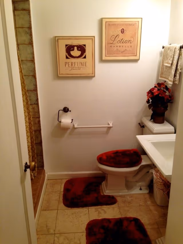 A small bathroom with beige tiled floor and walls. There is a toilet with a red fuzzy seat cover and matching red rugs on the floor. Above the toilet, there are two framed pictures on the wall, one labeled 'Perfume' and the other 'Lotion'. To the right of the toilet is a white sink with a countertop, and a vase with red flowers is placed on the toilet tank. A towel rack with a beige patterned towel is mounted on the wall above the flowers. On the left side, there is a shower area with a tiled wall and a shower curtain partially visible.