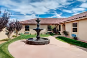 Outdoor courtyard area of Life Spire Assisted Living Rio Rancho featuring a central multi-tiered water fountain surrounded by a circular paved walkway, green grass, and beige stucco building walls under a partly cloudy sky.