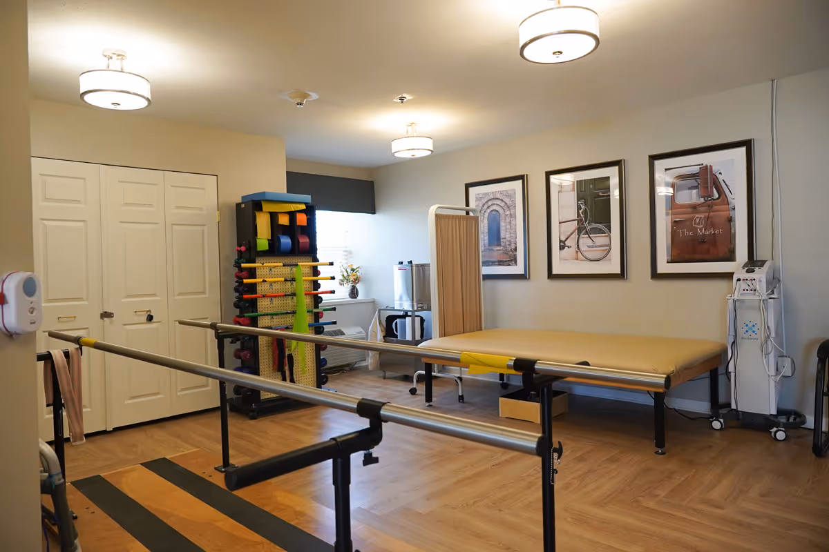 A physical therapy room with parallel bars for walking exercises, a padded treatment table, a rack holding various exercise bands and weights, and framed pictures on the wall. The room has wood flooring, soft lighting from ceiling fixtures, and a small window with a dark valance.