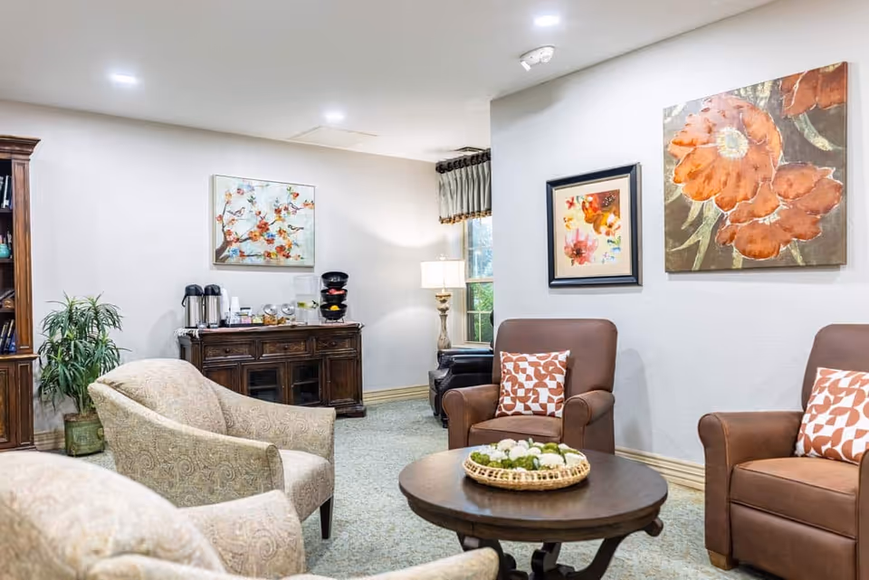 Comfortable common living area with upholstered chairs around a coffee table, artwork on the walls, and a refreshment cabinet.