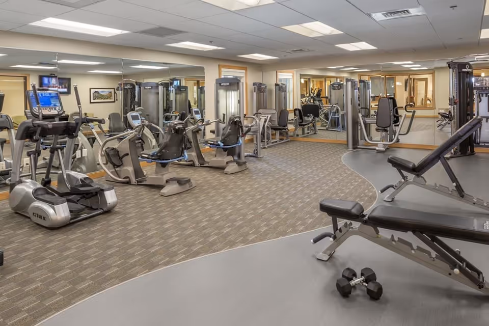 A well-lit fitness room with various exercise equipment including stationary bikes, weight machines, and benches. The room has a carpeted area and a mirrored wall reflecting the equipment and space.