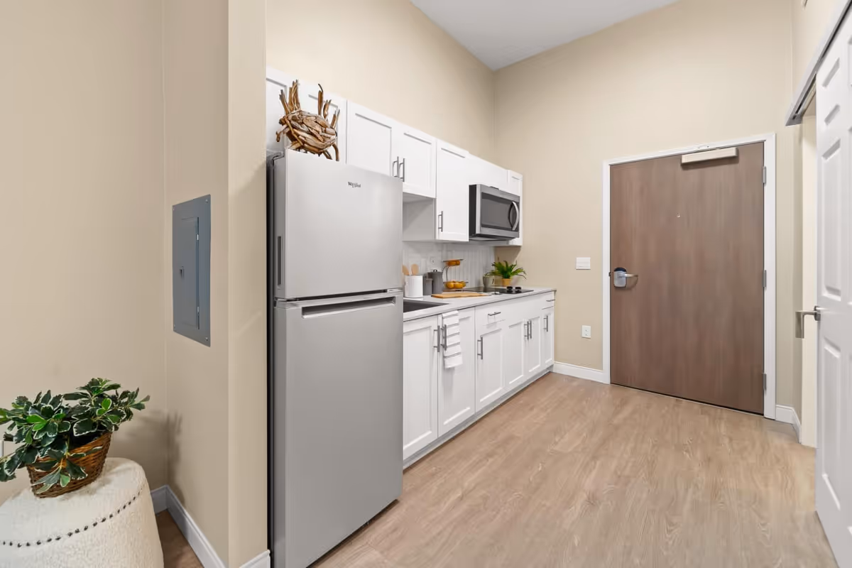 A small kitchen area with white cabinets, a stainless steel refrigerator, a microwave, and a countertop with kitchen utensils and a plant. The kitchen is adjacent to a wooden door and has light-colored walls and wood flooring.
