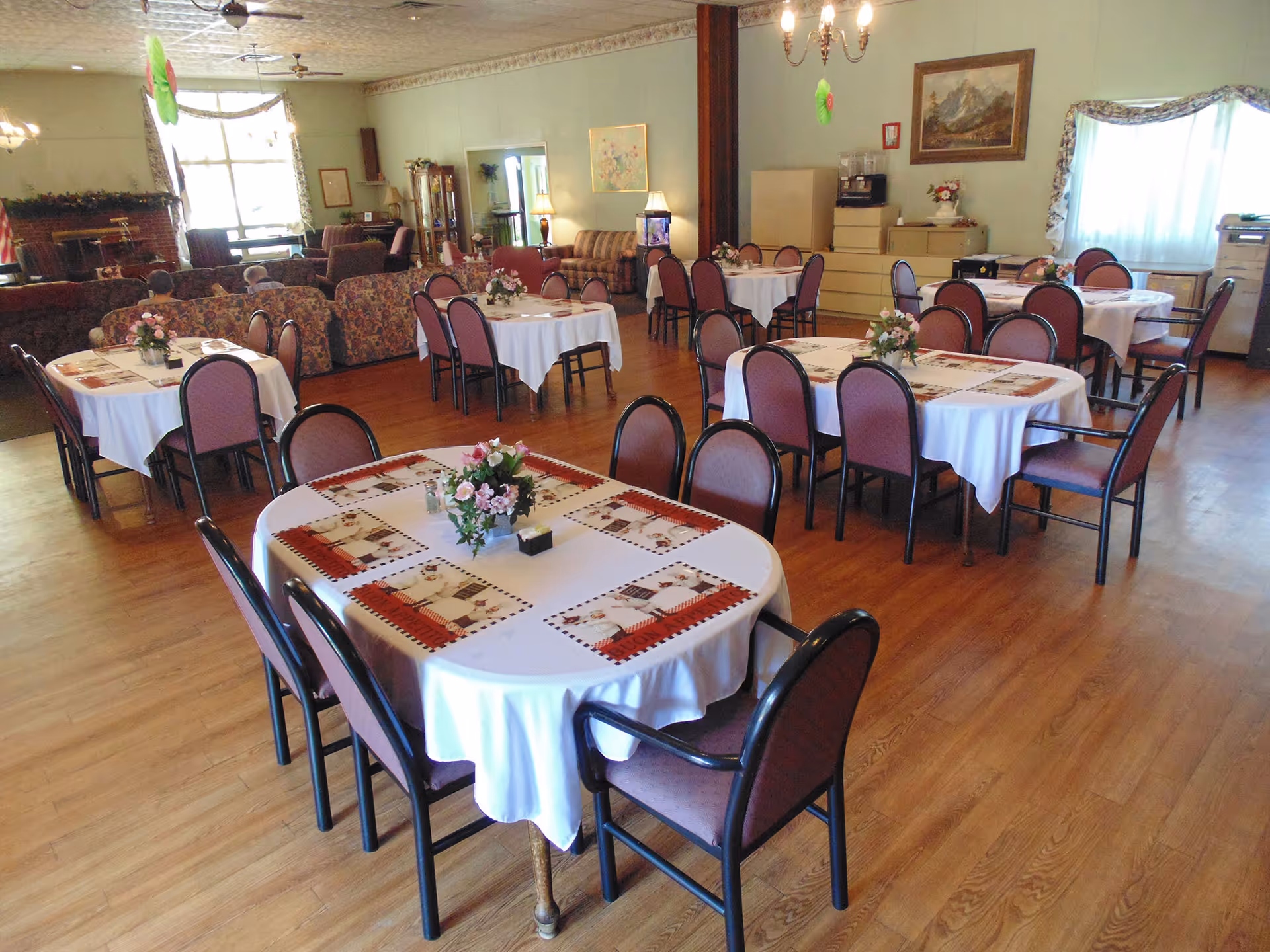 A dining room with several tables covered in white tablecloths and decorated with floral centerpieces. Each table is surrounded by chairs with pink upholstery. The room has wooden flooring, a chandelier, large windows with curtains, and a cozy seating area with couches and a fireplace in the background.