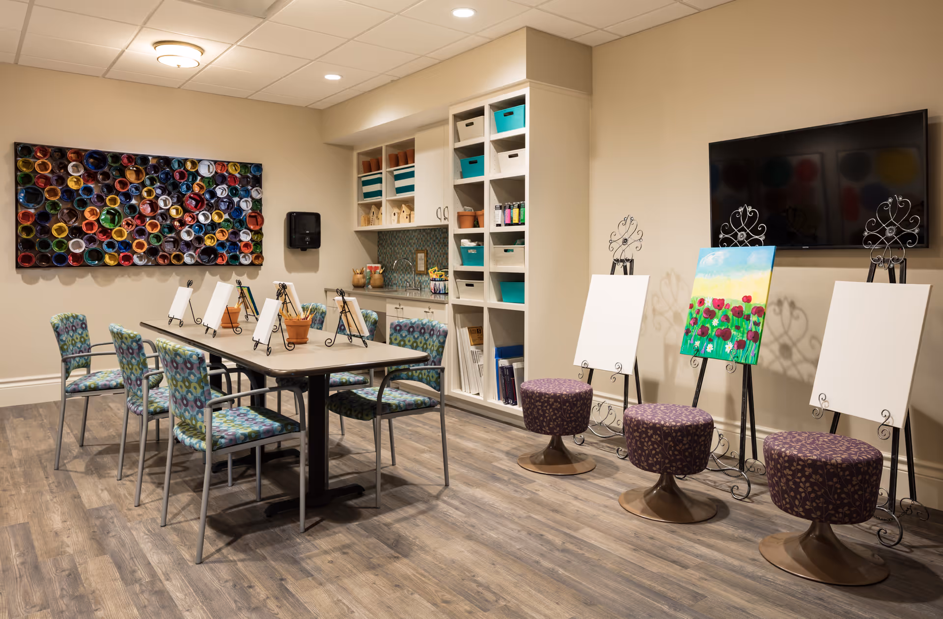 A well-lit art room with a long table surrounded by six patterned chairs. On the table are small easels holding blank canvases and art supplies. Against the wall are shelves with storage bins and art materials. Three additional easels with blank canvases and one painted canvas are positioned near a wall-mounted flat-screen TV. The room has wood flooring and a colorful circular wall art piece.