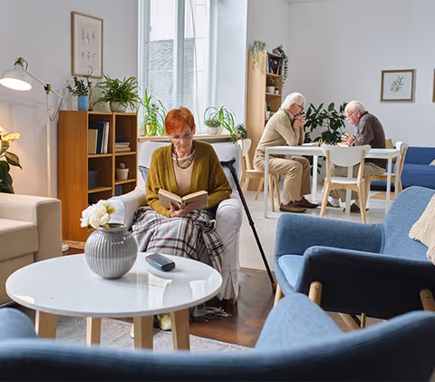 A cozy living room with a woman sitting in a white armchair reading a book, covered with a plaid blanket. In the background, two elderly men are sitting at a table engaged in conversation. The room is decorated with plants, framed pictures, and modern furniture including blue chairs and a white round coffee table with a vase of flowers.