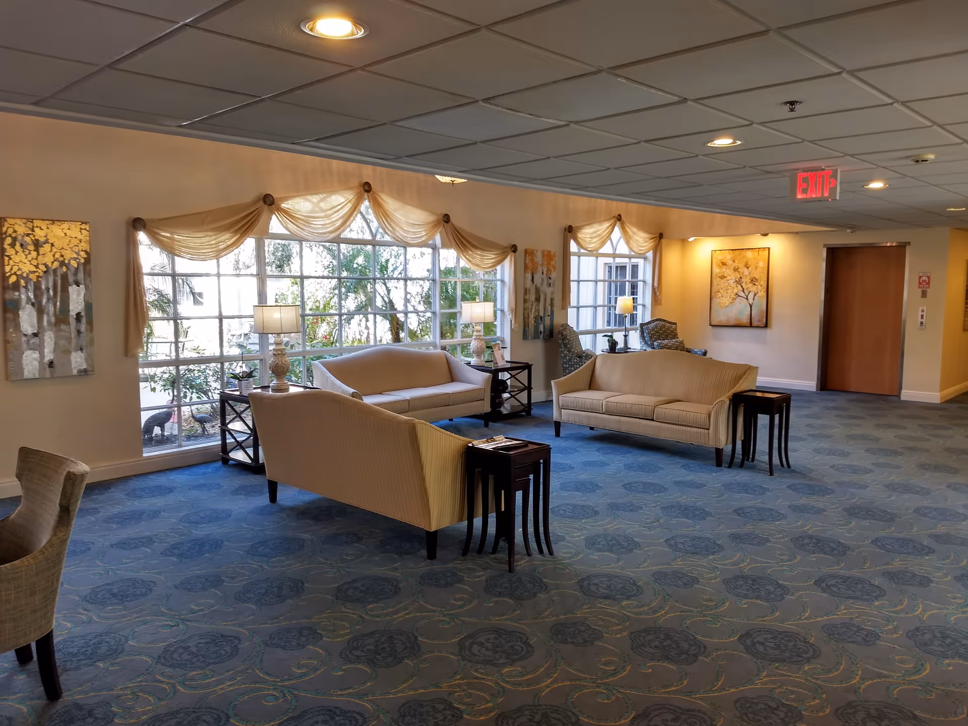 A spacious lounge area with two beige sofas facing each other, two side tables with lamps, two patterned armchairs near windows, and decorative paintings on the walls. Large windows with sheer curtains allow natural light to brighten the room, which has a blue patterned carpet and a ceiling with recessed lighting and an exit sign.