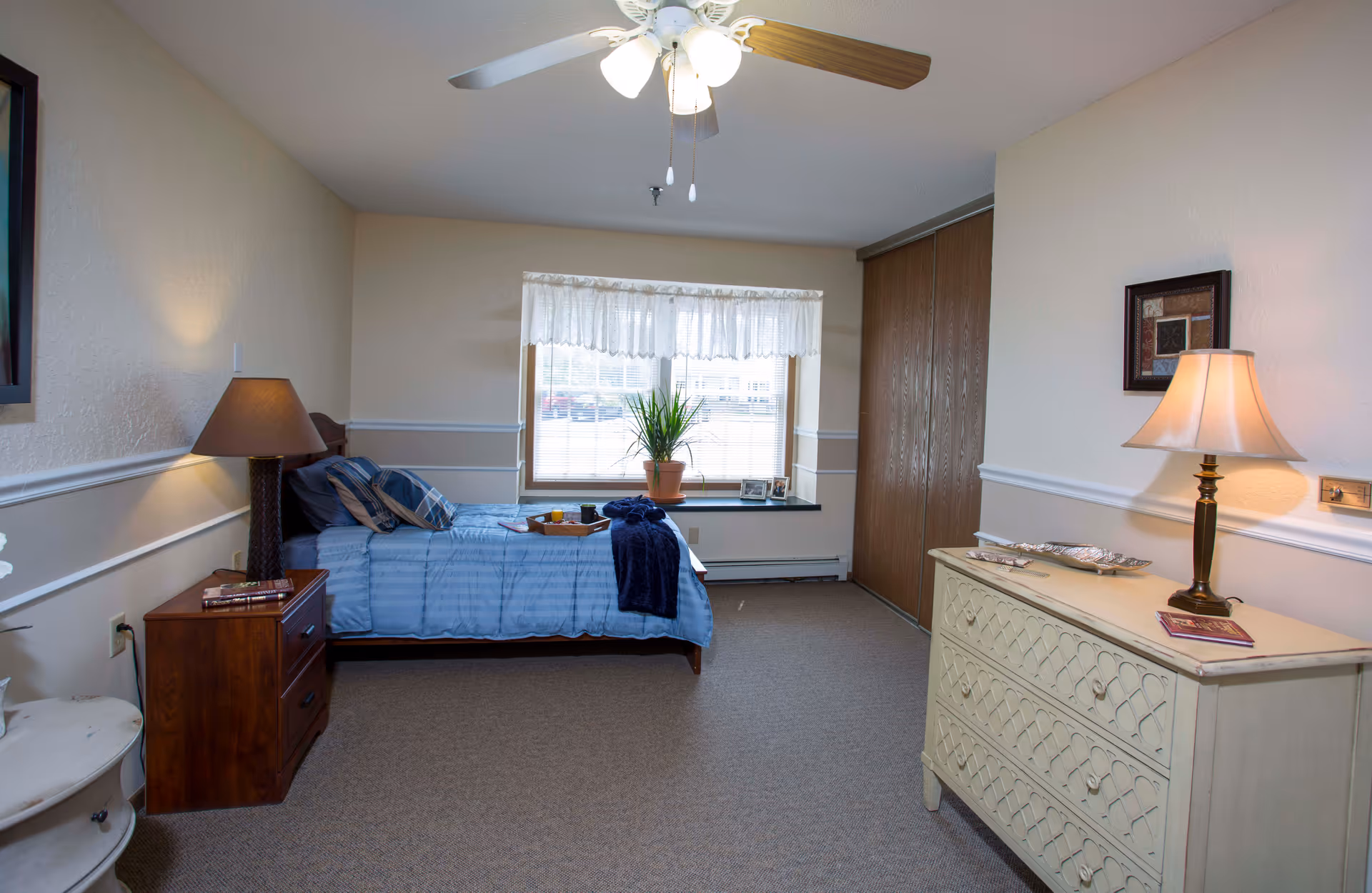 A bright bedroom with a single bed, bedside tables and lamps, a dresser, a window with a potted plant, and a ceiling fan.
