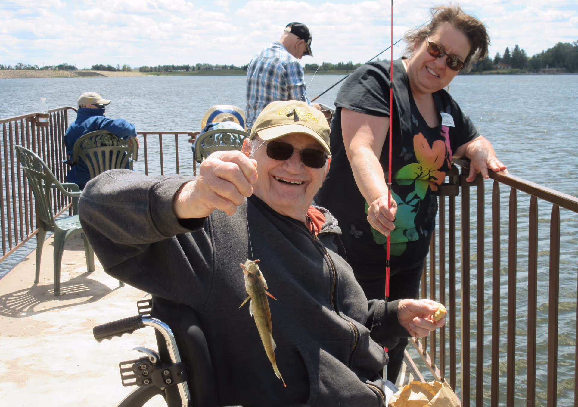 An elderly man in a wheelchair holding up a small fish he caught while fishing on a pier by a lake. A woman standing next to him is smiling and holding a fishing rod. Other people are fishing in the background on the pier under a partly cloudy sky.