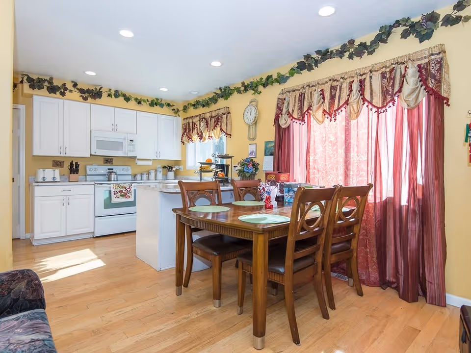 A bright kitchen and dining area with white cabinets, a white stove, microwave, and countertop appliances. A wooden dining table with four chairs is set with placemats and small decorations. The room has yellow walls, hardwood floors, and windows with red and gold curtains. Green leafy garlands decorate the top edges of the walls.