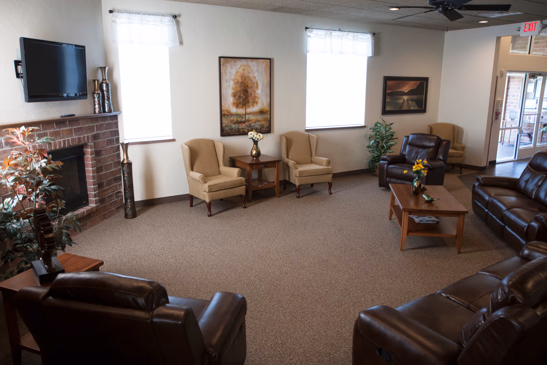 A cozy senior living facility common area with brown leather recliners and beige armchairs arranged around wooden coffee and side tables. The room features a brick fireplace with a mounted flat-screen TV above it, two windows with sheer white curtains, framed artwork on the walls, and decorative plants. An exit door with glass panels is visible in the background.