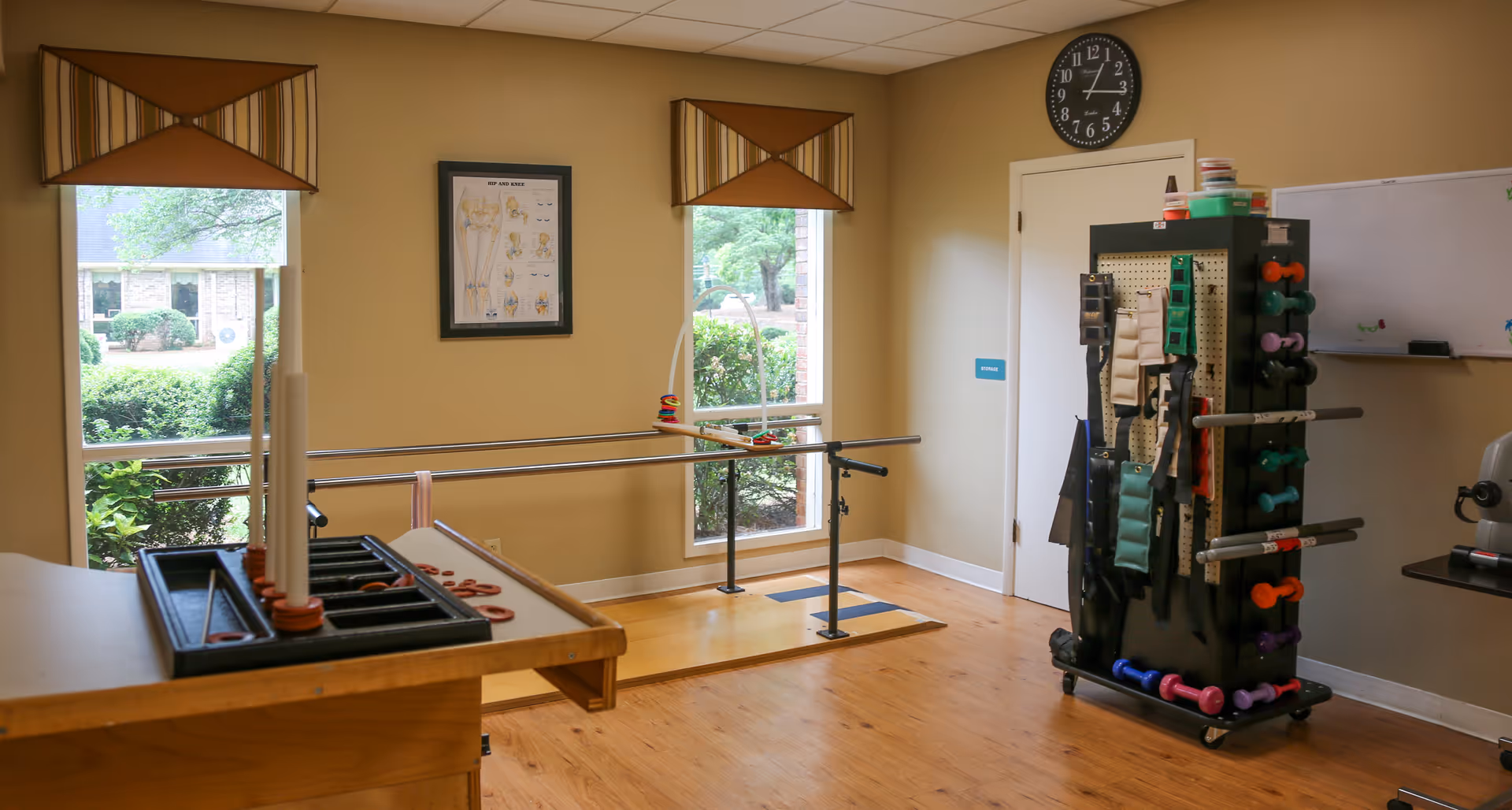 A rehabilitation or physical therapy room with wooden flooring, two large windows with striped valances, parallel bars for walking exercises, a rack holding various exercise bands and dumbbells, a wall clock, a whiteboard, and a wooden table with a ring toss game.