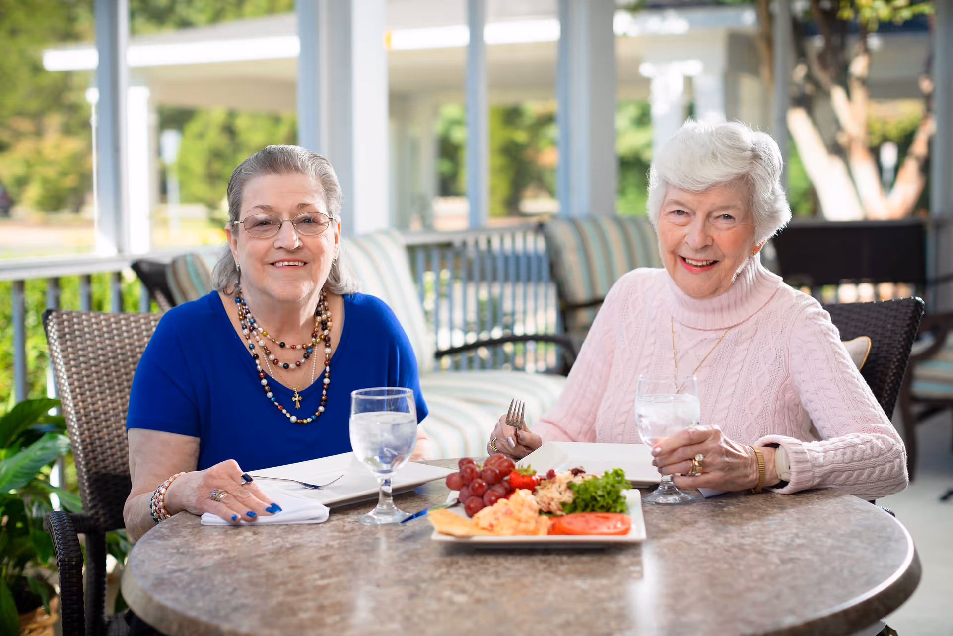 Two elderly women sitting at a round outdoor table with plates of food and glasses of water, smiling at the camera. The setting appears to be a patio area with cushioned chairs and greenery in the background.