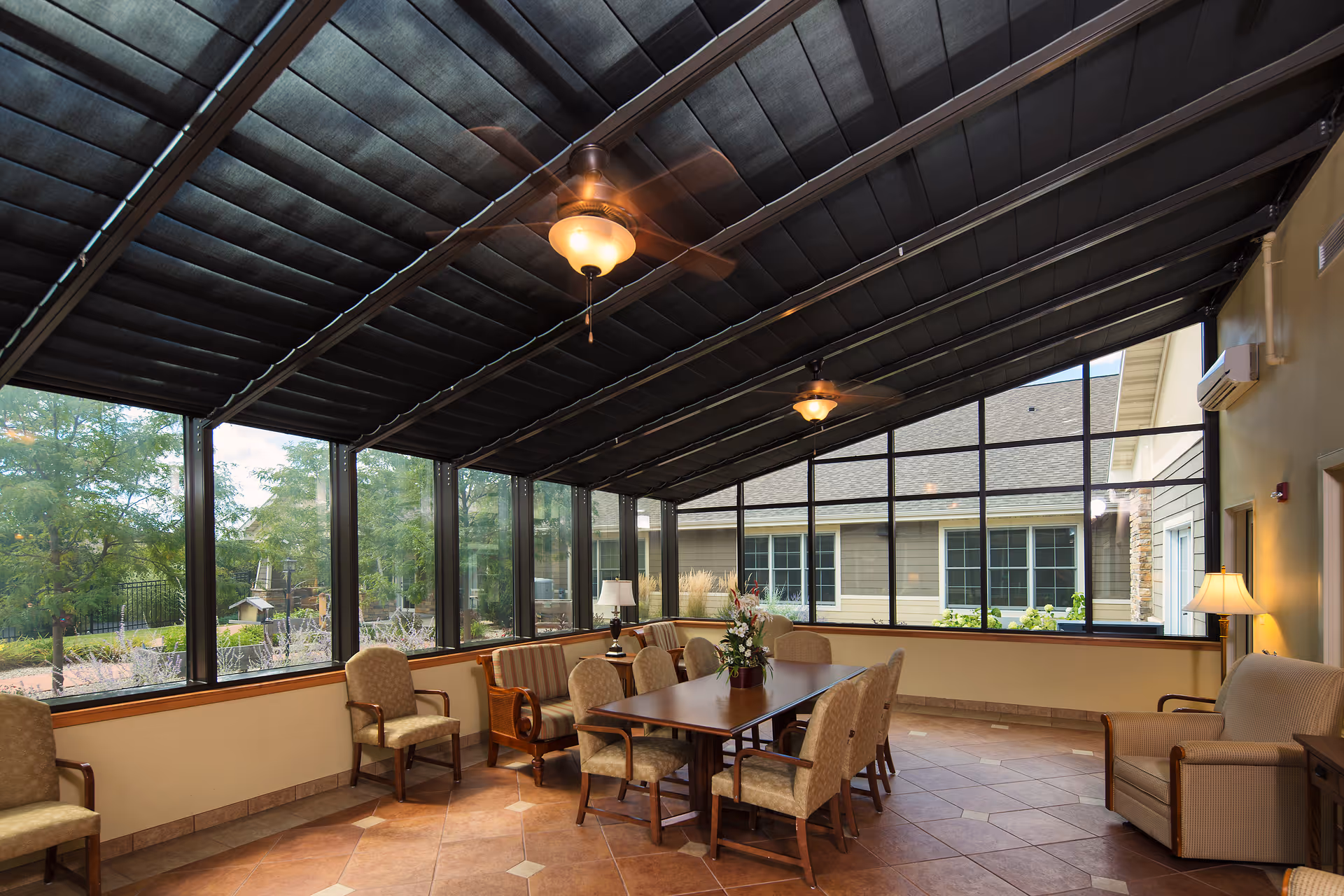 A sunroom or enclosed patio area with large glass windows and a slanted roof with ceiling fans. The room contains a rectangular wooden table surrounded by cushioned chairs and armchairs. There are lamps and a flower arrangement on the table, with a view of greenery and another building outside the windows.