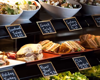 Deli display case showing sandwiches and bowls of salad with small chalkboard labels and prices.