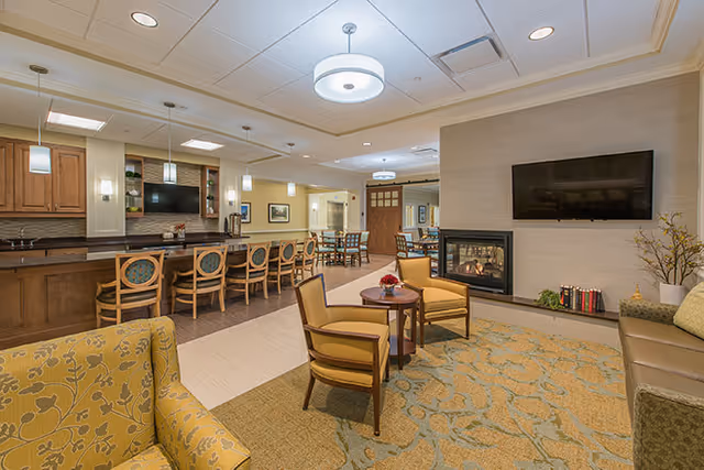 A spacious and well-lit common area in a senior living facility featuring a seating area with yellow patterned armchairs and a small round table, a wall-mounted flat-screen TV above a modern fireplace, and a kitchen area with a long counter and several high chairs. The room has warm lighting and neutral tones with decorative elements such as flowers and books.