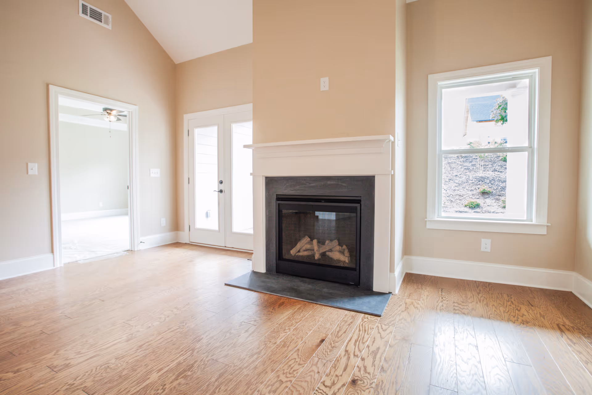 Bright room with wooden flooring featuring a modern fireplace with white mantel, a window showing an outdoor view, and a doorway leading to another room with a ceiling fan. There are also double glass doors leading outside.