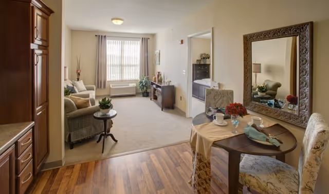 A cozy senior living apartment interior featuring a small dining table set for two with cups and napkins, a large ornate mirror on the wall, a living area with a sofa, armchair, side table with a plant, a TV stand with decorative items, and a window with vertical blinds letting in natural light.